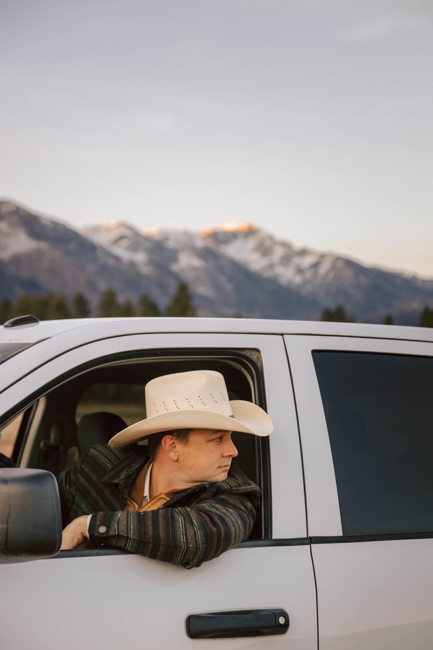 A man wearing Turner Hat Cowboy Canvas hat sitting in a vehicle with mountains in the background